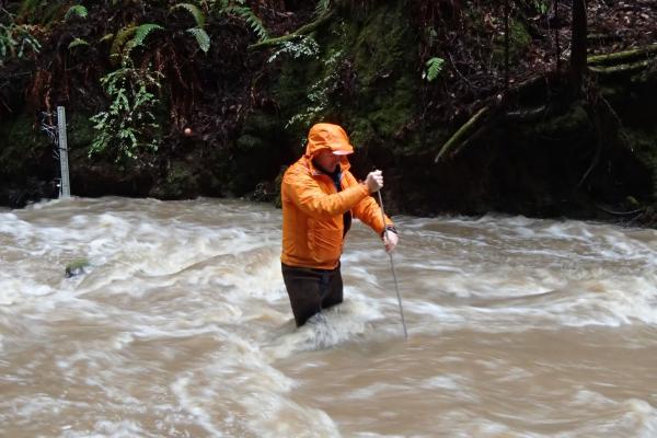 person in stream with brown water holding an instrument in the water. 