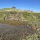 Pooled up water with a person standing downstream. Grassy hills and clear sky. 