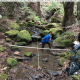 person in pool in a small creek holding a sediment probe. Two measureing tapes criscross the pool. picture has orientation information. 